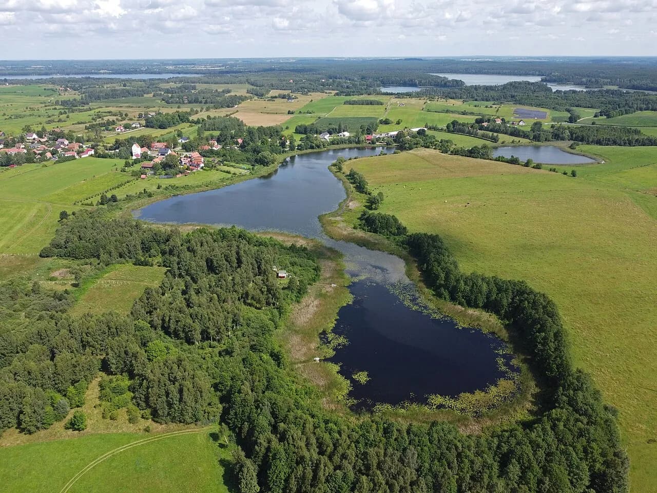 An aerial view of a winding lake surrounded by green farmland and forests, with a small village visible in the distance under a partly cloudy sky.