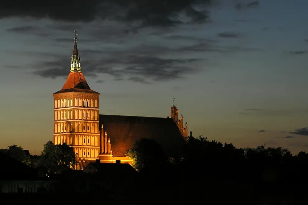 A church tower with a red roof glows with light against a dark blue evening sky.